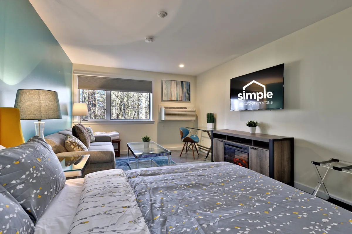 This image captures a light-filled, contemporary sleeping and lounging area featuring a queen bed with patterned bedding, a sofa and chairs for relaxing, a television with visible branding, and a window letting in natural light. The layout highlights both comfort and functionality in a cozy mountain retreat setting.