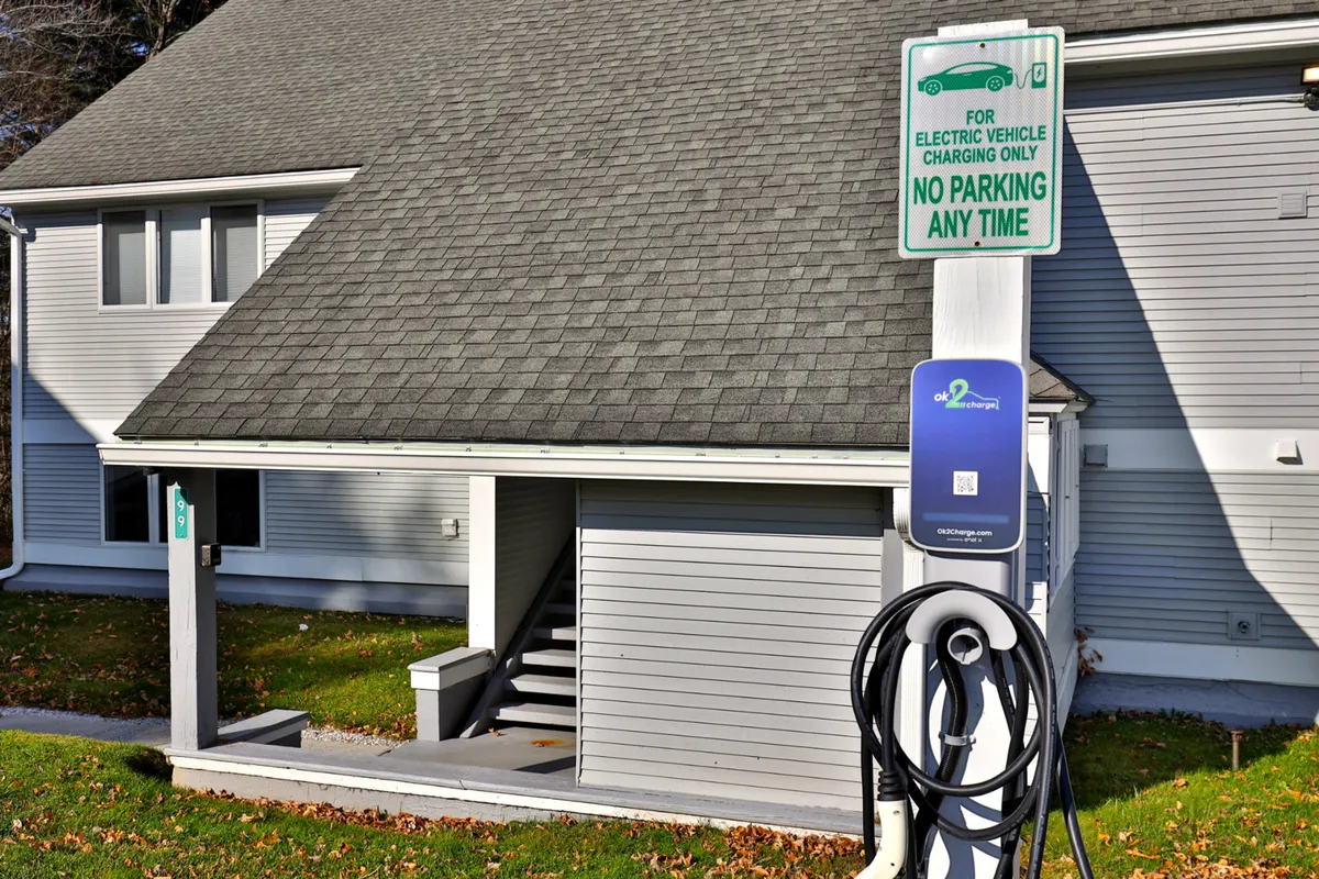 An exterior shot that highlights an electric vehicle charging station at the base of the building, stairs leading up to the entrance, and the building’s welcoming facade. The surrounding greenery and clear view of the parking area reinforce convenience and eco-friendly amenities.