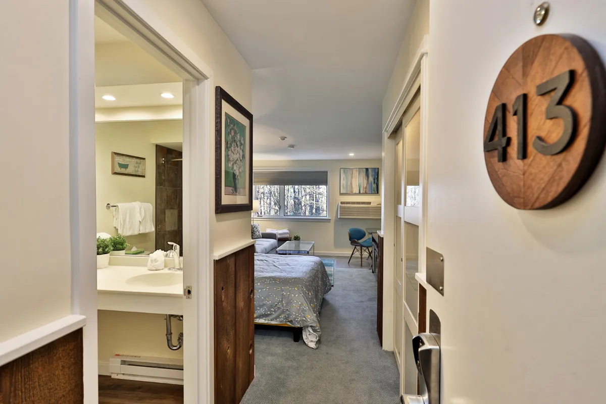 The image captures a bedroom scene with a patterned bed cover, a visible bathroom vanity, and a door marked with the room number. The setting is cozy and bright, evoking a sense of privacy and personal space.
