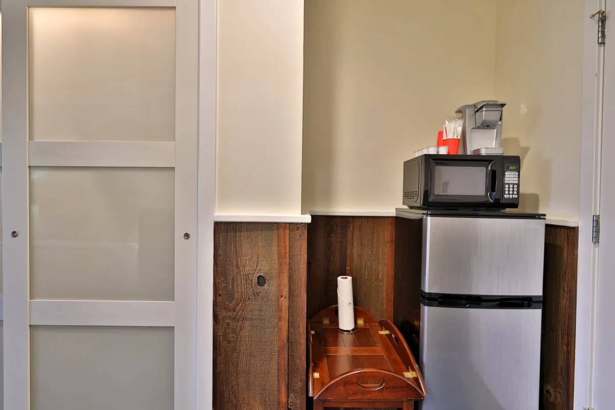 This image shows a kitchenette area with a refrigerator, microwave, and a wooden table, all well-lit and arranged for convenience. The setup clearly features essential appliances for preparing quick meals or snacks, with ample counter space and an inviting atmosphere.