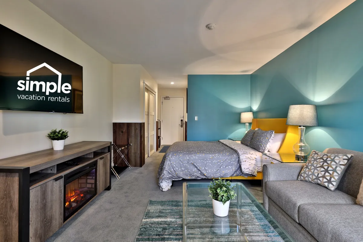 This image features a modern living space with a bed, yellow headboard, stylish sofa with decorative cushions, a TV with branding, and a coffee table with a potted plant. The overall feel is comfortable and well-appointed, with plenty of space for relaxation or work.