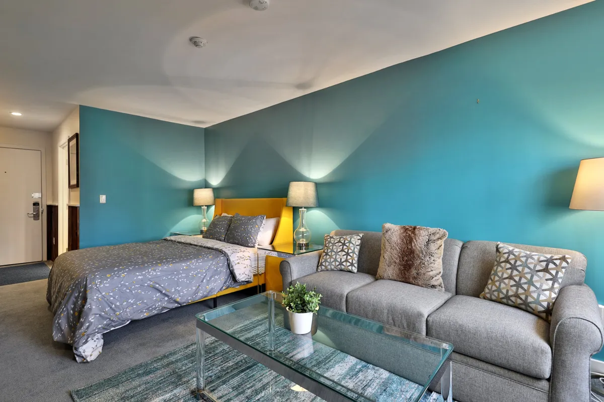 A cozy corner of the condo featuring a bed with a yellow headboard, a comfortable gray sofa, and a sleek glass coffee table, set up for both lounging and light social gatherings. Decorative pillows add a homey touch.
