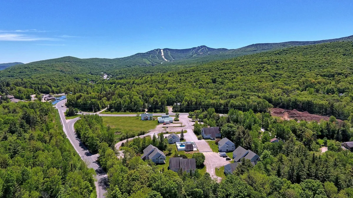 This exterior image captures a scenic view of the mountains, green trees, residential buildings, and a road leading toward the slopes. The setting is peaceful, with clear sightlines to the rugged terrain that becomes a playground for winter sports enthusiasts. The environment hints at its seasonal transformation, ideal for activities like skiing, snowboarding, and après-ski adventures.