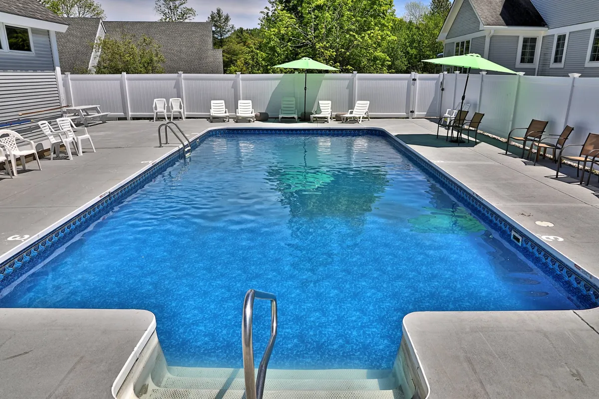 This cheerful exterior image focuses on the swimming pool area, highlighting inviting lounge chairs, green umbrellas, and the surrounding trees. The setup is perfect for sunbathing, socializing, or unwinding after a day of outdoor fun, capturing the lively yet relaxing summer atmosphere.