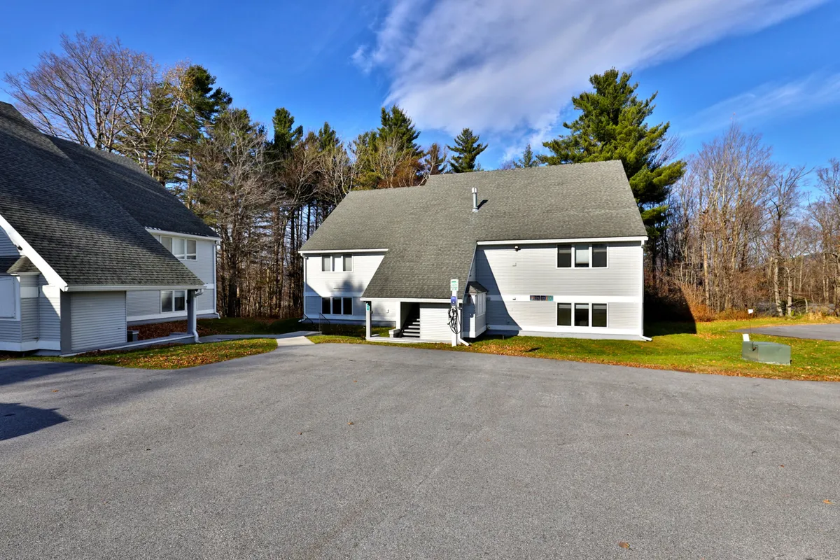 This tranquil exterior scene displays a two-story building surrounded by trees and a paved parking area, suggesting easy arrivals and departures regardless of the season. The natural setting evokes thoughts of seasonal activities and après-ski relaxation nearby.