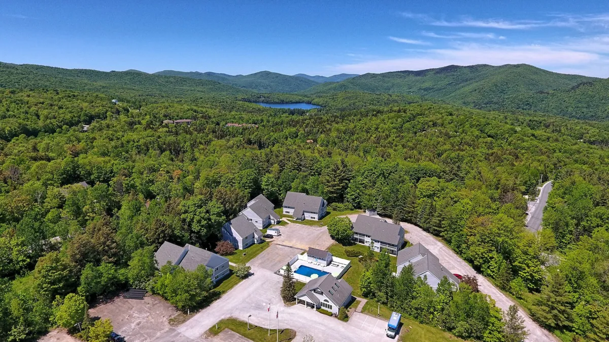 This exterior image features a sparkling swimming pool surrounded by a cluster of condo buildings and lush greenery, with the majestic Vermont mountain range rising in the background. The setting feels integrated with nature, inviting guests to swim, relax, or enjoy outdoor activities against a scenic backdrop.