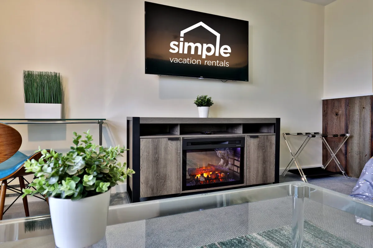 A warm and inviting living area with a glowing electric fireplace, a large TV with visible branding, decorative plants, and a glass coffee table. The space is perfect for relaxing on a cozy evening, watching TV, or sharing stories by the fire.