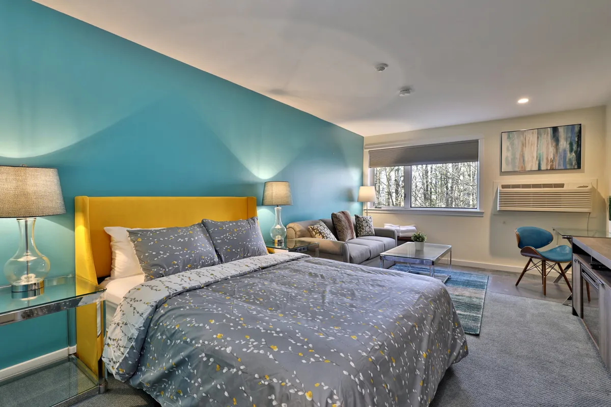 The image showcases a bright and inviting studio condo interior, centered around a cheerful yellow bed frame against a bold blue accent wall. A comfortable sofa seating area sits nearby, complemented by modern decorative artwork. There's a window offering natural light, creating a cozy, relaxing corner ideal for enjoying morning coffee or unwinding after a day outdoors.