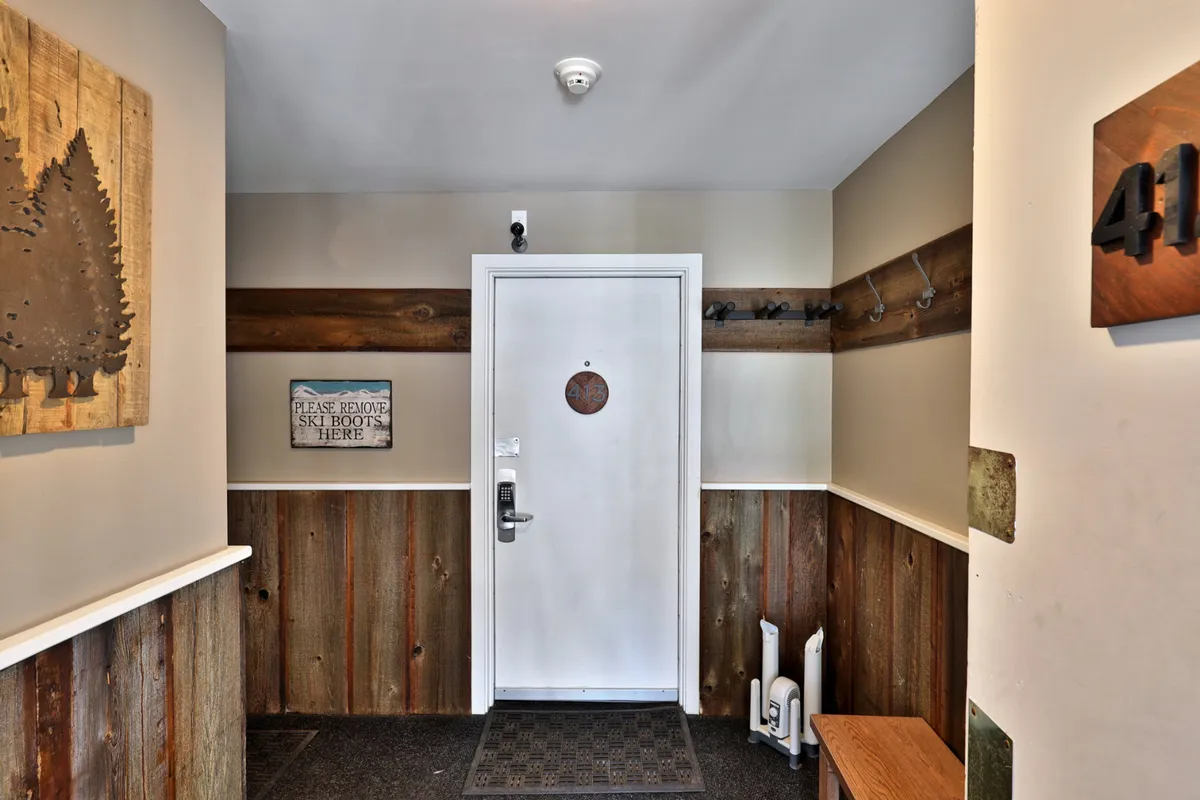 A welcoming view of the studio's entryway featuring a front door, wooden wall art, a coatrack for jackets and ski gear, and a cheerful welcome mat. The space exudes warmth and practicality, inviting guests to easily drop off outdoor equipment and transition inside after a day of adventure.