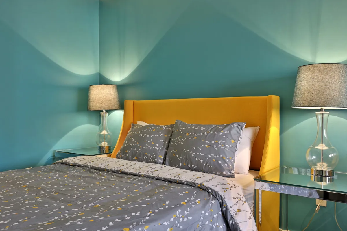 A cozy interior room with a yellow upholstered headboard, gray and yellow patterned bedding, and silver lamps. The atmosphere is vibrant and inviting, ideal for a restful break or productive work session.